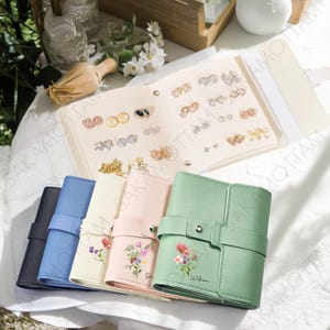 以下が含まれることがあります： さまざまなイヤリングが展示された開いたジュエリーオーガナイザーと、カラフルな花の装飾が施されたジュエリーケースの列。 ケースは、ネイビー、ブルー、クリーム、ピンク、グリーンの色合いです。 オーガナイザーとケースは白い表面にあります。