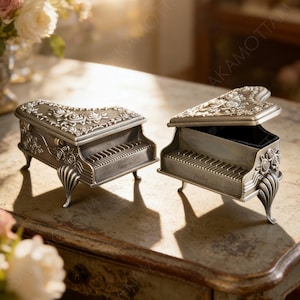 以下が含まれることがあります： 2つの銀色のピアノ型ジュエリーボックス。1つのボックスは開いており、黒い内側が見えます。どちらのボックスにも花柄とスクロールワークのディテールがあり、「Remember」と「Harley」という名前がキーボードエリアに刻まれています。