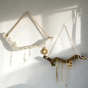 以下が含まれることがあります： ロープで吊るされた枝で作られた2つの素朴なジュエリーオーガナイザー。1つは白く塗装され、もう1つは天然木です。どちらも、ジュエリーを掛けるための小さな金色のペグを備えています。さまざまなネックレスとイヤリングが表示されています。