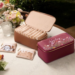 以下が含まれることがあります： ジッパー開閉式の茶色の革製ジュエリーボックス。複数のコンパートメントが現れるように開いています。「Harley」という名前と花の模様が入ったバーガンディ色のジュエリーボックスと、ジュエリーと時計用の小さなポーチも写っています。