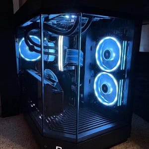 Puede incluir: Una caja de ordenador negra con un panel lateral de vidrio, que muestra un PC personalizado con iluminación LED azul. La caja tiene un diseño elegante y moderno y cuenta con varios ventiladores con luces LED azules.