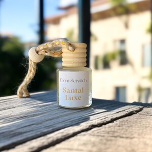 May include: A small glass bottle with a wooden cap and a rope loop. The label reads "From Scratch Santal Luxe." The bottle sits on a weathered wooden surface, with a blurred background of buildings and sky.