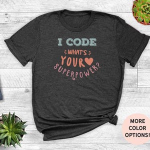 May include: Dark grey t-shirt featuring the text "I CODE WHAT'S YOUR SUPERPOWER?" in light blue and pink, with a heart symbol. This casual t-shirt is suitable for everyday wear.
