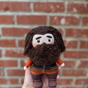 May include: Crocheted amigurumi doll of a bearded man with brown hair, wearing a brown, orange, and black outfit. The doll is holding a small, white heart.