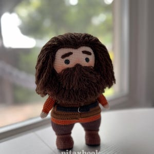 May include: A crocheted plush toy of a character with brown hair and a beard, wearing a brown, orange, and black outfit. The toy is standing on a white surface. The text "pita&hearts;hook" is visible on the surface.