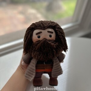 May include: Crocheted amigurumi doll of a wizard with a brown beard and brown hair. The doll is wearing a brown coat, brown pants, and brown boots. The doll is holding a wand in its right hand.