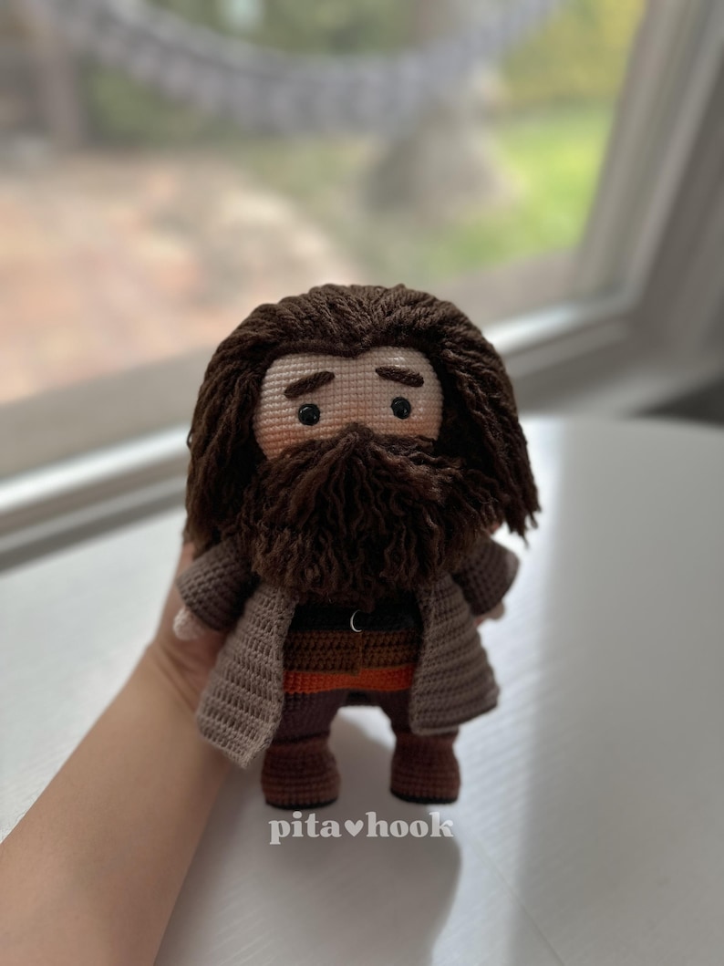 May include: Crocheted amigurumi doll of a wizard with a brown beard and brown hair. The doll is wearing a brown coat, brown pants, and brown boots. The doll is holding a wand in its right hand.