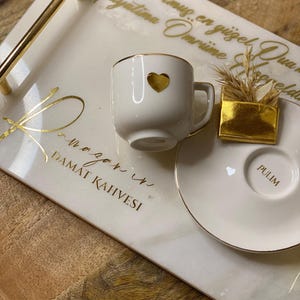 May include: A white ceramic coffee cup with a gold heart design, gold trim, and a matching saucer. The cup and saucer sit on a decorative tray with gold handles and text. A small gold box with wheat is also on the tray.