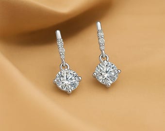 May include: A pair of silver drop earrings featuring a sparkling, round, clear gemstone. The earrings have a small decorative bar with smaller clear gemstones. The background is a warm, golden colour.