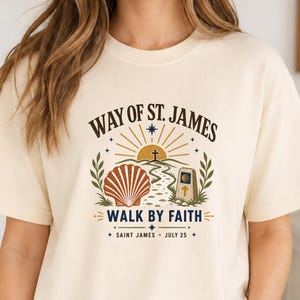 May include: Cream-colored t-shirt featuring a graphic design with the text "WAY OF ST. JAMES" above an illustration of a shell, path, and cross. The phrase "WALK BY FAITH" is below the illustration, with "SAINT JAMES JULY 25" at the bottom.
