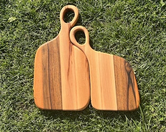 Op de afbeelding: Twee houten snijplanken met handvatten, een groter dan de andere, liggen op een groene grasmat. De planken zijn gemaakt van licht en donker hout, waardoor een gestreept patroon ontstaat.