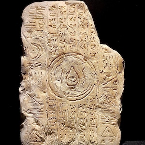 May include: A weathered, rectangular stone tablet with carved symbols and inscriptions. The beige-colored tablet features a central circular design with a stylized eye and teardrop. Various geometric shapes and lines are etched into the surface.