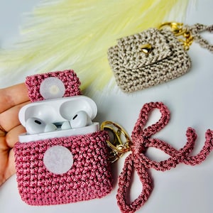 Op de afbeelding: Twee gehaakte AirPod-hoesjes met sleutelhangers. Een hoesje is roze met een witte cirkel en een roze lint-sleutelhanger. Het andere hoesje is beige met een gouden knoop en een gouden ketting-sleutelhanger.