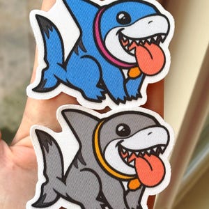 May include: Two embroidered iron-on patches of cartoon sharks with open mouths and tongues sticking out. The shark on top is blue with a pink collar, and the shark on the bottom is gray with a yellow collar.