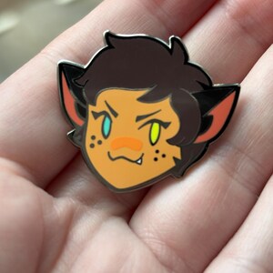 May include: A cartoon enamel pin featuring a character with brown hair, orange skin, and green and blue eyes. The character has cat ears and a small mouth with a fang showing. The pin is silver with black outlines.