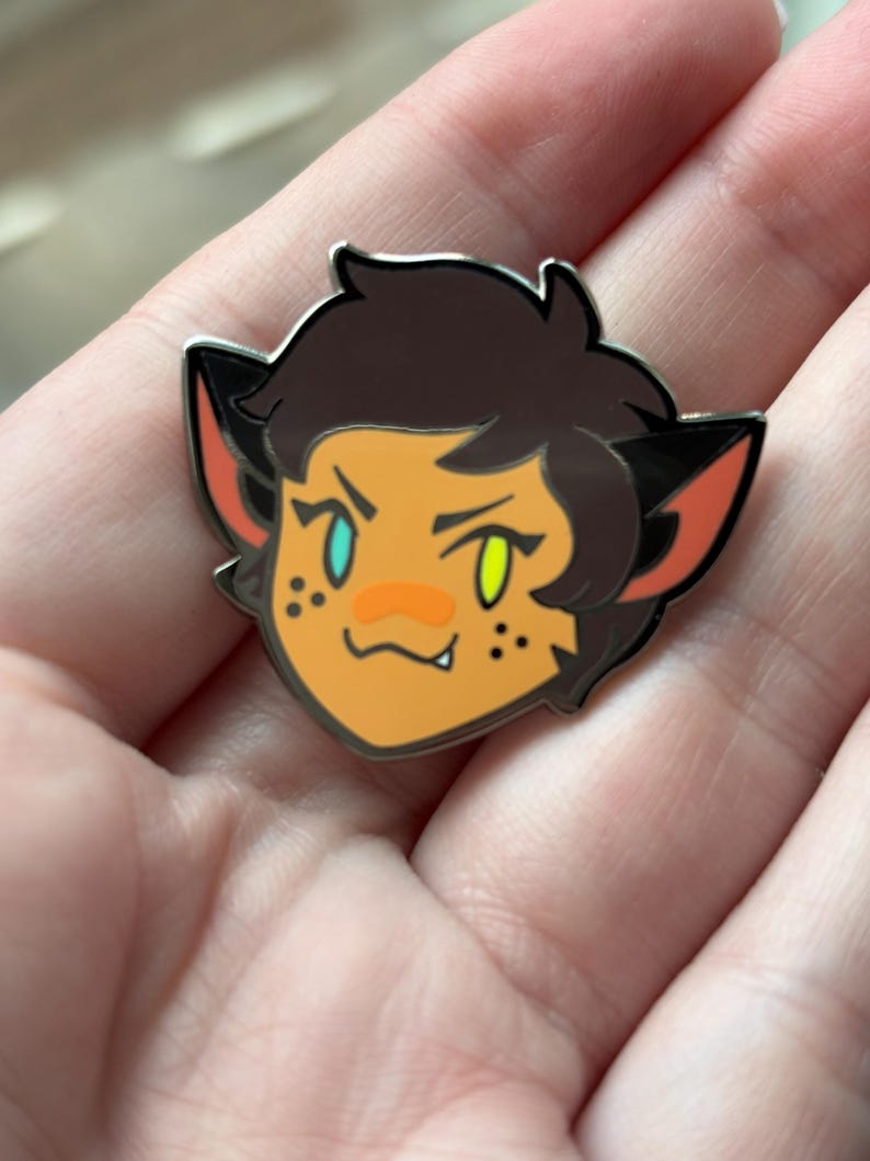 May include: A cartoon enamel pin featuring a character with brown hair, orange skin, and green and blue eyes. The character has cat ears and a small mouth with a fang showing. The pin is silver with black outlines.