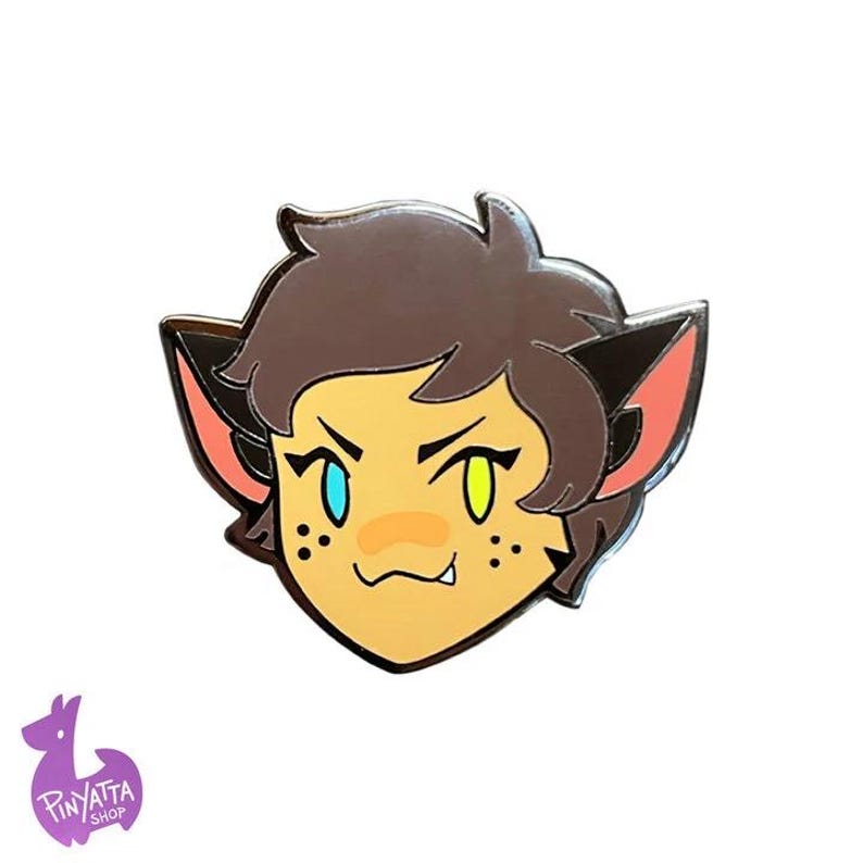 May include: A cartoon enamel pin featuring a cat-like character with brown hair, green and blue eyes, and a pink nose. The character has a mischievous expression and is wearing a pink collar.