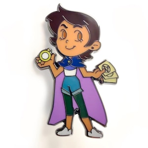 Puede incluir: Pin esmaltado que representa un personaje de dibujos animados con cabello castaño, que lleva una capa y pantalones cortos azules, y sostiene un objeto dorado y cartas. El pin tiene un contorno plateado sobre un fondo blanco.