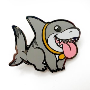 May include: A gray cartoon shark with a yellow collar and a pink tongue sticking out. The shark is smiling and has big eyes. The pin is made of metal.