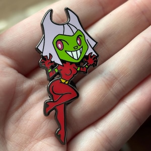 Può includere: Uno spillo in smalto verde e rosso con un personaggio dei cartoni animati con un ampio sorriso, capelli bianchi e vestiti rossi. Il personaggio è in posa con una gamba sollevata e entrambe le braccia alzate.