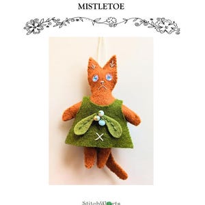 以下が含まれることがあります： 手作りのフェルトクリスマス猫オーナメント。オレンジ色の猫は青い目を持ち、緑色のドレスを着ています。画像には「Christmas Cat Embroidered Felt Pattern Mistletoe」と書かれています。