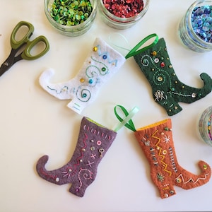 May include: Four felt elf shoe ornaments in white, dark green, purple, and orange, each decorated with embroidery, sequins, and small charms. A pair of green-handled scissors and four glass jars filled with colorful sequins are nearby.