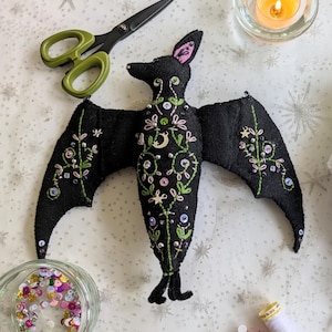 May include: A black felt bat plush toy featuring intricate green vine embroidery, pink floral stitching, and small metallic star and moon charms. The bat is decorated with various beads and buttons, with pink inner ears and a pointed tail.