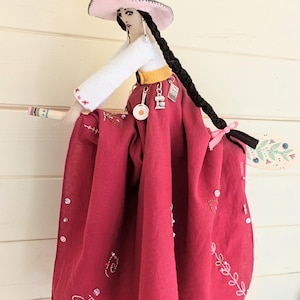 May include: A fabric doll dressed as a witch, wearing a grey and pink pointed hat, a white long-sleeved top, and a long dark red skirt with embroidered floral patterns. The doll has long black braided hair, silver charms, and holds a wooden broom.