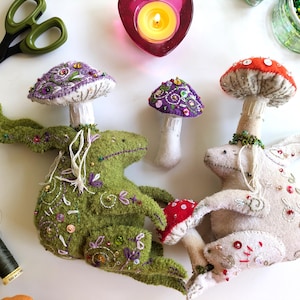 May include: Two plush rabbit figures with mushroom caps on their heads, one green and one beige, decorated with intricate embroidery, beads, and sequins. A smaller purple mushroom figure sits between them, accompanied by sewing tools and thread.