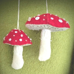 Può includere: Due ornamenti a forma di fungo realizzati in feltro. Ognuno presenta un cappello rosso vivace decorato con accenti circolari bianchi e perline di vetro trasparenti, fissato su un gambo spesso color bianco sporco. Fili bianchi servono per appenderli.
