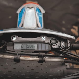 Puede incluir: Primer plano del salpicadero de una motocicleta, con una pantalla digital que muestra números. La motocicleta tiene un manillar negro y un carenado delantero blanco y azul. El salpicadero incluye un velocímetro y un interruptor de encendido.