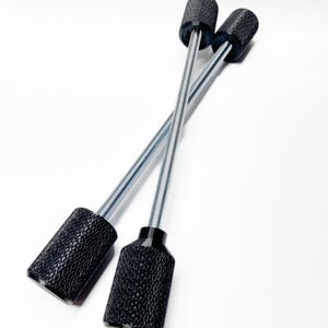 May include: Two black, textured cylindrical objects with silver metal rods extending from them, crossing each other diagonally against a white background. The cylindrical objects have a diamond pattern.