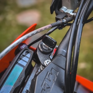 Pode incluir: Close-up da área do guiador de uma motocicleta, com um interruptor de ignição preto com uma chave inserida. A imagem destaca o logotipo da KTM, um visor digital e vários cabos e controlos pretos. A motocicleta tem detalhes em laranja.