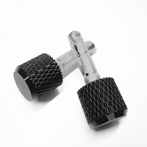 May include: Two silver-toned metal cufflinks with black, textured cylindrical ends. The cufflinks are connected by a silver bar. The textured ends have a repeating pattern. The background is white.