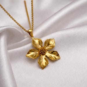 May include: A gold-colored flower pendant necklace. The pendant is a detailed, five-petal flower with a textured surface. The necklace chain is also gold and hangs on a white, silky background.