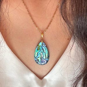 May include: A gold-colored chain necklace with a teardrop-shaped pendant. The pendant features a vibrant, iridescent abalone shell design with shades of blue, green, and purple. The necklace is worn against a white top.