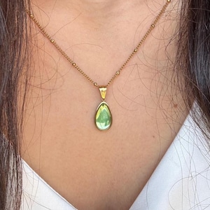 May include: A gold-colored necklace with a pear-shaped, light green gemstone pendant. The pendant is framed in gold and hangs from a delicate chain. The necklace is worn against a white top.