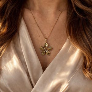 Puede incluir: Un collar dorado con un colgante de flor. El colgante tiene p&eacute;talos detallados en forma de hoja y cuelga de una fina cadena. El colgante floral es de color oro y verde, sobre una tela color crema. Es una joya.