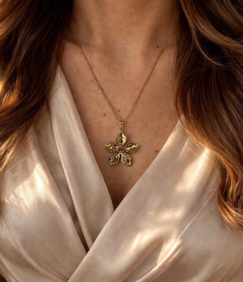 Puede incluir: Un collar dorado con un colgante de flor. El colgante tiene p&eacute;talos detallados en forma de hoja y cuelga de una fina cadena. El colgante floral es de color oro y verde, sobre una tela color crema. Es una joya.