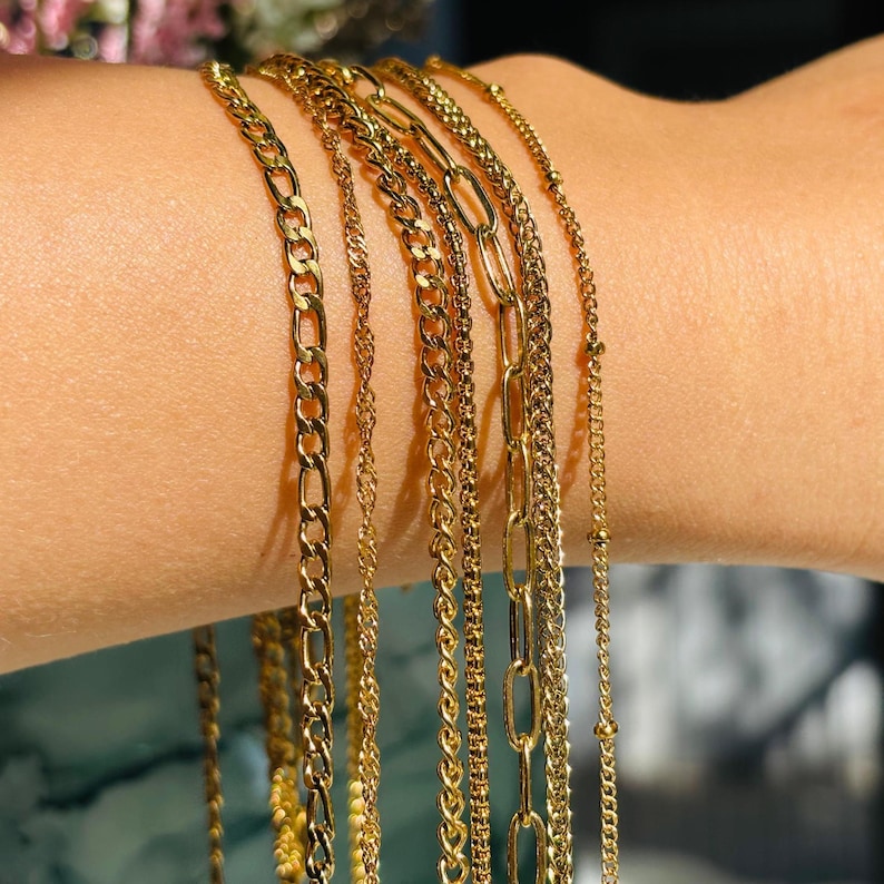May include: A collection of gold-colored chain bracelets in various styles. The bracelets include curb chains, rope chains, and delicate beaded chains. The jewelry is displayed on a person's arm, showcasing the different textures and designs.