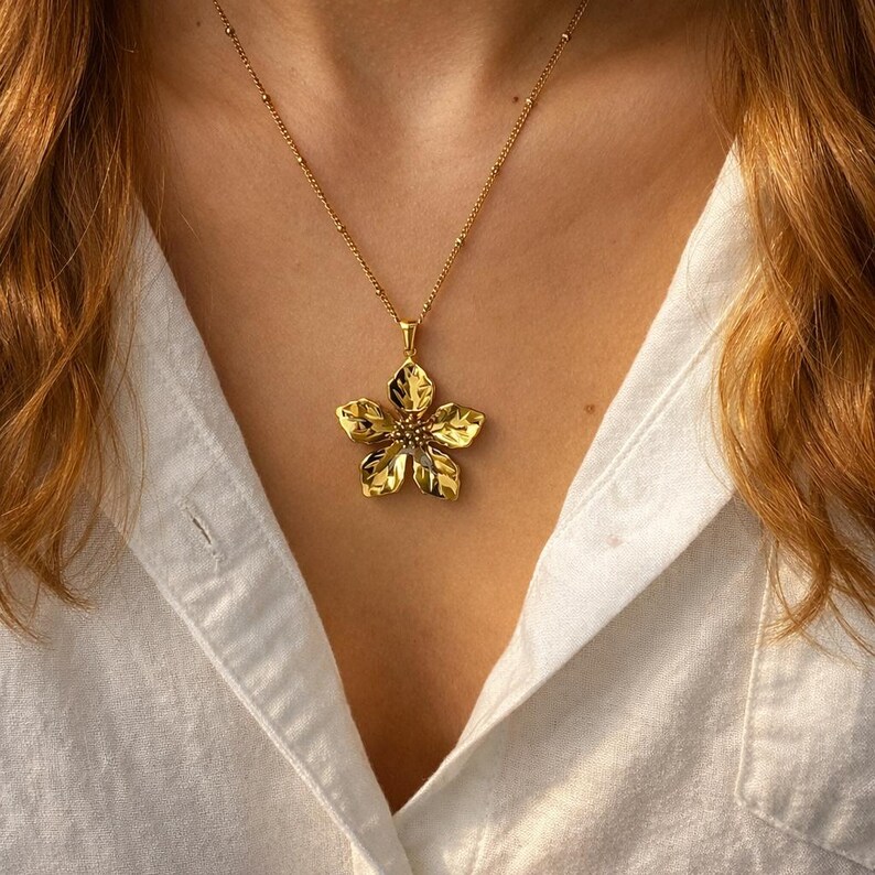 Puede incluir: Un collar dorado con un colgante de flor con un dise&ntilde;o detallado de p&eacute;talos. El collar se lleva sobre una camisa blanca, mostrando la intrincada artesan&iacute;a y el estilo elegante de la joya. La cadena es delicada y complementa el colgante floral.