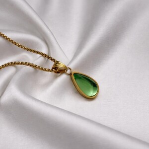 May include: A gold-toned necklace with a teardrop-shaped green pendant. The pendant is set in a gold frame and hangs from a delicate chain. The necklace rests on a white, silky fabric.
