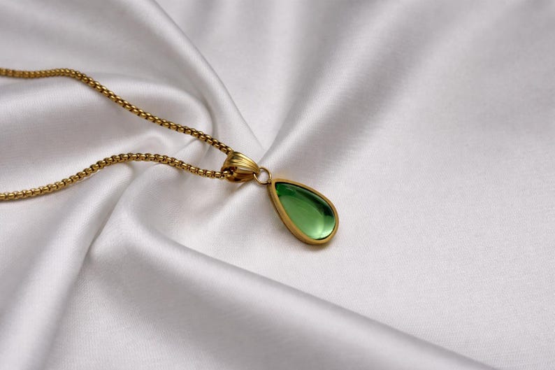 May include: A gold-toned necklace with a teardrop-shaped green pendant. The pendant is set in a gold frame and hangs from a delicate chain. The necklace rests on a white, silky fabric.