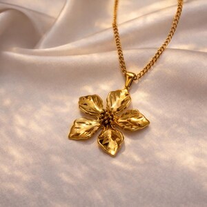 Puede incluir: Un collar dorado con un colgante de flor. El colgante tiene cinco p&eacute;talos y una superficie texturizada. La cadena del collar tambi&eacute;n es dorada y descansa sobre una tela de sat&eacute;n blanca.