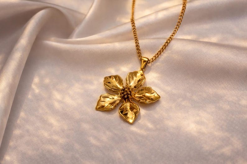 Puede incluir: Un collar dorado con un colgante de flor. El colgante tiene cinco p&eacute;talos y una superficie texturizada. La cadena del collar tambi&eacute;n es dorada y descansa sobre una tela de sat&eacute;n blanca.