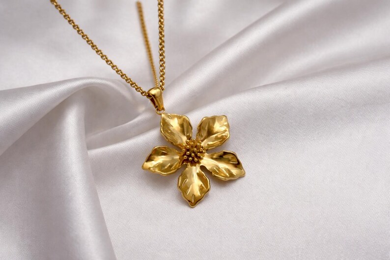 May include: A gold-colored flower pendant necklace. The pendant is a detailed, five-petal flower with a textured surface. The necklace chain is also gold and hangs on a white, silky background.