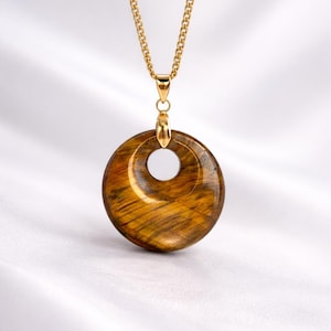 May include: A gold-colored chain necklace with a pendant. The pendant is a circular, polished tiger's eye stone with a hole in the center. The stone has brown and gold tones. The necklace is displayed on a white background.