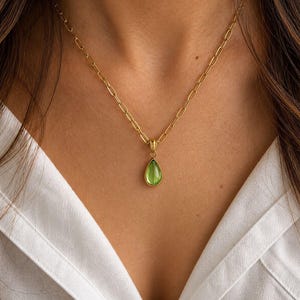 May include: A gold-colored necklace with a pear-shaped, light green gemstone pendant. The pendant hangs from a delicate chain. The necklace is worn against a white collared shirt.