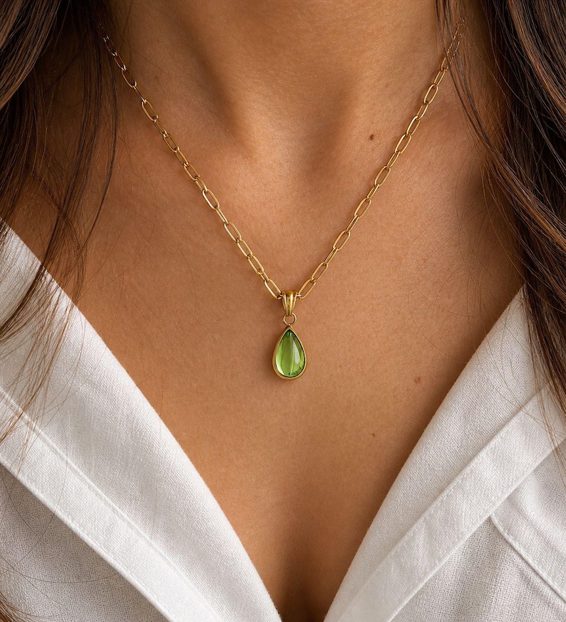 May include: A gold-colored necklace with a pear-shaped, light green gemstone pendant. The pendant hangs from a delicate chain. The necklace is worn against a white collared shirt.
