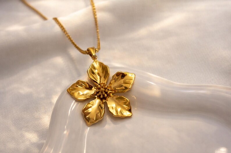 May include: A gold-colored necklace featuring a large, detailed flower pendant. The pendant has five petals and a textured surface, suspended from a delicate chain. The necklace is displayed on a white surface.